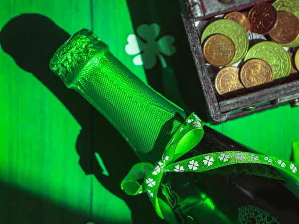 One bottle of green beer with a ribbon with paper leaves clover and a chest of coins on a green. — Stock Photo, Image