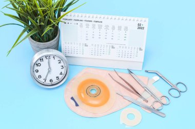 Saat yedinci saat, Şubat 2022 takvimi, cerrahi aletler, kolostomi torbası ve saksıda bir çiçek açık mavi arka planda, yakın planda. Kolon kanseri konsepti, ameliyat ve ameliyat planlaması.