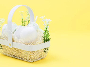 Beyaz metal hasır sepetli saman, yapay bahar çiçekleri ve solda sarı arkaplanda paskalya yumurtaları. Sağ tarafta metin için yer var, yan görünüm yakın. Mutlu Paskalya konsepti, bayrak ve kurusıkı.