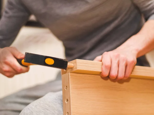 The hands of a Caucasian young man hammer a wooden nail into the headboard with a yellow hammer, sitting on the floor in his room, close-up side view. Concept of assembling furniture, shopping, at