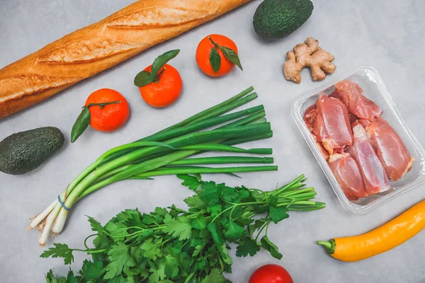 Fresh produce: french baguette, avocado, tangerines, fresh parsley, young green onions, tomatoes, peppers, ginger root and raw chicken thighs lie diagonally on a light gray cement background, close-up
