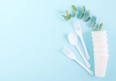 Plastik beyaz gözlüklü, kaşıklı, çatallı ve yeşil dallı solda fotokopi alanı olan açık mavi bir sıranın üzerinde duruyor. Ekoloji konsepti ve tek kullanımlık çatal bıçak..