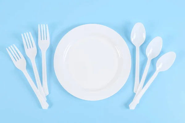 Empty plastic plate with forks and spoons on the sides on a blue background, flat lay close-up. Eco-friendly concept, disposable cutlery and ecology day.