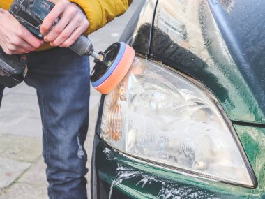 Kafkasyalı genç bir adamın elleri, süngerli bir matkapla matkabı deliyor ve arabasının farını deterjan sabunuyla parlatıyor. Yakın plan, araba yıkama ve araba farlarıyla temizlik kavramı..