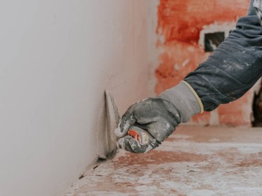 A young caucasian guy in a gray long-sleeve t-shirt and gloves holds a spatula with white putty and plasters the wall from below while sitting on the floor in the apartment, close-up side view. The concept of apartment and house renovation, wall plas