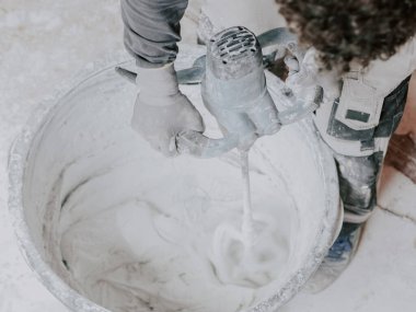 A young caucasian guy a plasterer in a gray long-sleeved T-shirt, gloves and work pants holds a construction electric mixer and stirs dry putty with water preparing it for work, close-up side view. The concept of apartment and house renovation, wall 