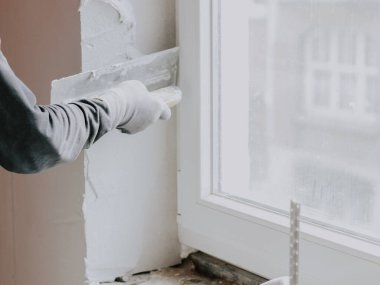 Gri tişörtlü, kollu bir adamın eli pencere aralığına küçük bir spatula ile yeni macun sürüyor. Evlerin ve dairelerin tamiri, duvarların sıvalanması, sıva hizmetleri..