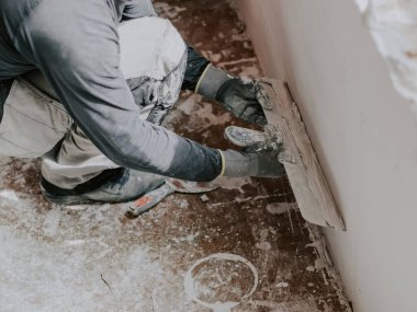 Gri tişörtlü ve kıvırcık kahverengi saçlı beyaz bir adam pencerenin altındaki duvarda büyük bir spatulayla çömelmiş taze macunu uyguluyor. Daire ve ev tadilatı, duvar sıvası, alçı.