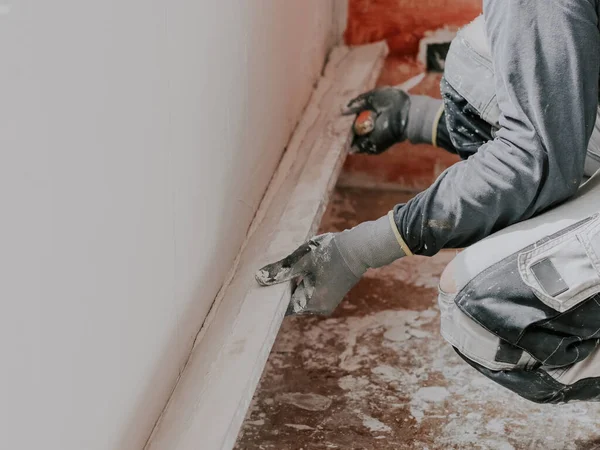 Young caucasian guy plasterer in gray long sleeve t-shirts, construction gloves holds a wooden rule and levels a freshly puttyed wall while squatting on the floor, close-up side view. The concept of apartment and house renovation, wall plastering, pl