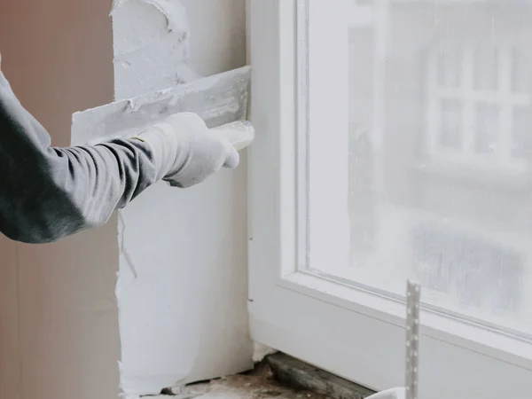 The hand of a guy in a gray T-shirt with sleeves applies fresh putty with a small spatula to the window opening, side view close-up. The concept of repairing apartments and houses, plastering walls, plastering services, at home.