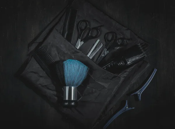 Scissors, combs, hair clipper, brush and clips with an apron lie on a black homemade wooden background in the center, flat lay close-up. The concept of male hairdressing, dark style.