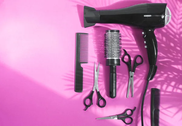 Scissors, hair dryer, hair straighteners and combs lie to the right in a semicircle on a pink background with copy space spit with shadow from palm branches, flat lay close up.Hairdressing concept.