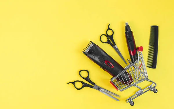 Scissors, combs and hair and ear clippers, nose clippers lie in a mini cart on the right on a yellow background with copy space on the left, flat close-up. Male hairdressing and shopping concept.