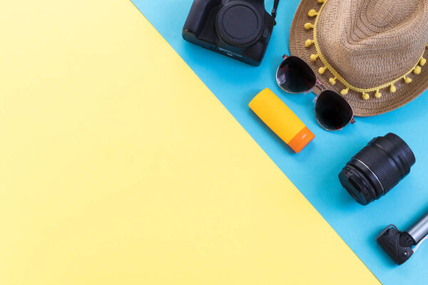 Camera, straw hat, sunscreen, sunglasses and lens on right side on yellow and blue background with copy space on left side, flat lay closeup. Summer concept, blogger.