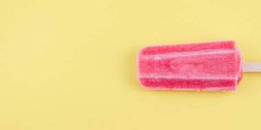 Ahşap bir çubukta ahududu şerbeti sağda sarı bir zemin üzerinde duruyor solda fotokopi alanı var, düz yakın plan. Dondurma konsepti, şerbet, yaz, bayrak, minimalizm.