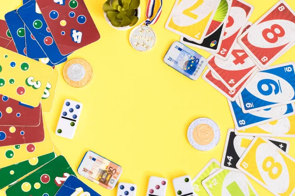 Old,dirty,colorful game cards ligretto uno,dominoes,chocolate money and winner medal lie in a round frame on a yellow background with copy space in the center,flat lay closeup.Summer board games concept.