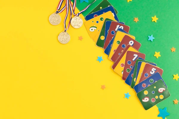 Old, dirty, colorful ligretto game cards, three winner medals and felt stars lie on the right on a green-yellow background with copy space on the left, flat lay close-up. Summer board games concept.