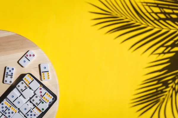 White dominoes with colorful dots in a metal box lie on a round wooden spinning board on the left on a yellow background with a shadow of a palm branch and copy space on the right, flat lay close-up. Concept summer board game.