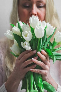 Beyaz lalelerden oluşan bir buket, beyaz bir buket, güzel bir manikürlü güzel beyaz bir sarışın kadının avuçlarında, çiçeklere nazikçe bakıyor, beyaz bir duvara bakıyor, derinliği...