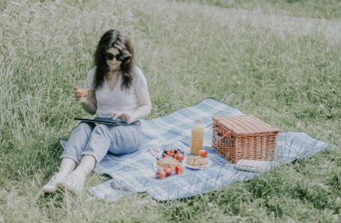 Güneş gözlüklü, kahverengi saçlı, kot pantolonlu ve tişörtlü genç beyaz bir kız elinde hasır bir sepetle yerde oturuyor, tabakta meyveler ve elinde taze bir tabletle sosyal ağlara bakıyor.