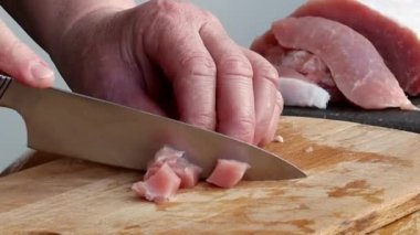 Hands of a senior caucasian woman cutting a pork fillet with a knife into small cubes on a cutting board at the table,close-up side view.Concept cooking.