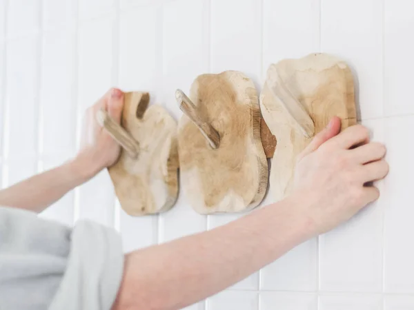 The hands of a young caucasian guy in a gray fleece robe with rolled up sleeves hangs a new wooden stylish and eco-friendly hanger on the wall with white tiles in the bathroom, close-up from below
