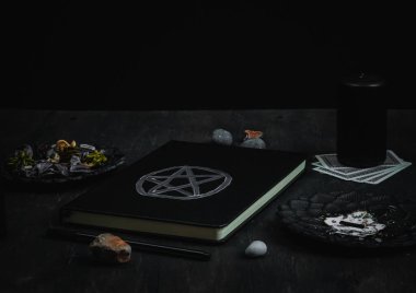 Sihirli beyaz sembollü siyah deri bir kitap, siyah mumlar, siyah metal tabaklar, kurutulmuş çiçekler, ritüel sopaları, tarot kartları, siyah ahşap arka planda bir kalem ve taşlar, yakın plan yan görüş. Esrarengiz konsept, karanlık stil. Deri kitap.