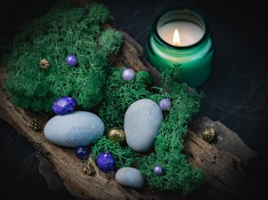 Lilac, golden natural stone lies in the bark of a tree with green moss, gray stones and a burning candle in a glass jar on a black wooden background, close-up side view with depth of field. Esoteric