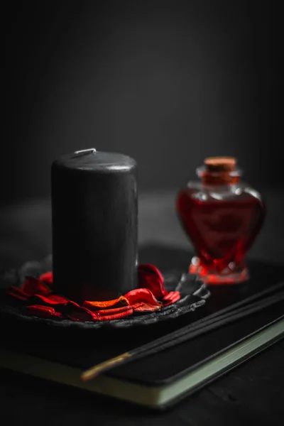 A black candle with a red potion in a heart shape bottle and ritual sticks lie on a magic book in the center on a black wooden background, close-up side view. Esoteric concept, dark style.