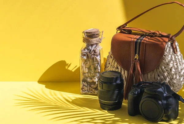 Camera, shells in a glass bottle and lens on the right on a yellow background with a shadow of palm branches, side view close-up. Summer concept, blogger.