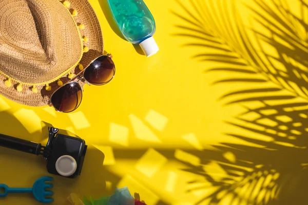 Camera, sunblock, sunglasses and a straw hat on the right on a yellow background with palm branches shadow, sun glare and copy space on the left, flat lay close-up. Summer concept, blogger.