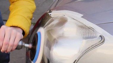 The hands of a young caucasian man in a yellow jacket are polishing with a detergent and a polishing machine the front headlight of a car, side view close-up in slow motion.Concept headlight polishing,polishing service.