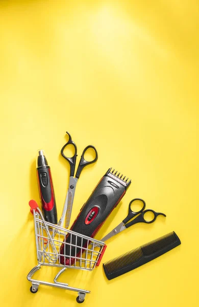 Scissors, combs and hair and ear clippers, nose clippers lie in a mini cart from below on a yellow background with copy space on top, flat lay close-up. Men\'s barbershop and shopping concept.