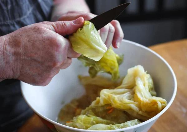 The hands of a senior woman hold a leaf of sauerkraut and cut off the rough part with a knife, preparing it for cabbage rolls, close-up side view. Concept step by step instruction, cooking home, traditional recipes, national cuisine, cabbage rolls, d