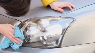 The hands of a young caucasian man in a yellow jacket are wiping the front headlight of a green car with a blue rag with detergent, side view close-up in slow motion.Concept Headlight polishing, polisher services.