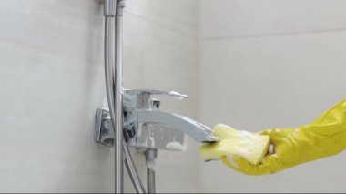 The hands of a caucasian young woman in yellow gloves rub a dirty faucet in the bathroom with a sponge and detergent, side view close-up in slow motion. Cleaning the bathroom concept.