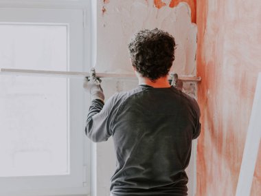 A young caucasian guy in a gray t-shirt with sleeves and curly brown hair from the back smoothes fresh putty on the wall near the window with a wooden rule, close-up side view. The concept of repairing apartments and houses, plastering walls, plaster