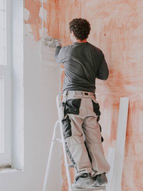 A young caucasian guy in a gray t-shirt with sleeves, work pants and curly brown hair applies fresh putty with a small spatula on the wall near the window, standing on a stepladder, close-up side view. The concept of apartment and house renovation, w