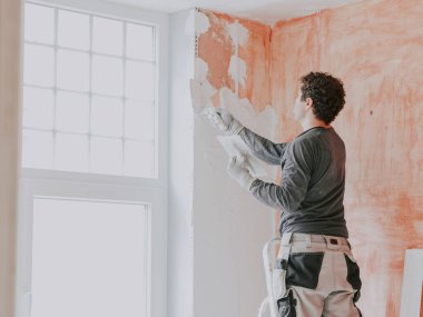 A young caucasian guy in a gray t-shirt with sleeves, work pants and curly brown hair applies fresh putty with a small spatula on the wall near the window, standing on a stepladder, close-up side view. The concept of apartment and house renovation, w