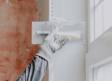 One hand in a construction glove, a plasterer in a gray T-shirt with sleeves, applies fresh putty on a window opening with a spatula, close-up side view. The concept of apartment and house renovation, wall plastering, plastering services, at home.