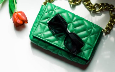 Stylish women's trendy green quilted bag with a large gold chain, a red tulip in it and sunglasses lie on a white table with shadows from the window, flat lay close-up. Fashionable accessories concept.
