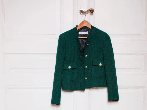 Stylish women\'s trendy green jacket with gold large buttons hangs on a wooden coat hanger over the doorknob of an antique white door with little copy space, close-up side view. The concept of fashion accessories.