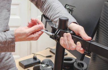 The hands of a young caucasian guy without a face twists a nut on a bracket with a wrench, installing a computer monitor on it against the background of blurry doors and a desk in the room, side view close-up. The concept of repair at home.