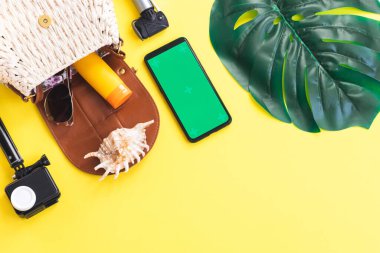 One camera, straw bag, sunglasses, suntan lotion, shell, monstera leaf and smartphone with green screen on left on yellow background with copy space on right, flat lay close-up. Summer concept, summer blogger, summer time.