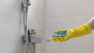The hands of a caucasian young woman in yellow gloves rub a dirty faucet in the bathroom with a sponge and detergent, side view close-up in slow motion. Cleaning the bathroom concept.