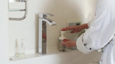 Hands of a caucasian young woman in a white bathrobe squeezing out liquid soap from a handwashing bottle in the bathroom, close-up side view in slow motion. Concept body care,healthy lifestyle.