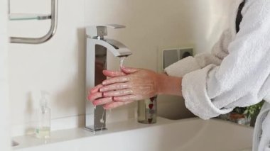 Caucasian young woman in a white bathrobe rinses her hands under a stream of clean water flowing from a faucet in the bathroom, close-up side view in slow motion. Concept body care,healthy lifestyle.