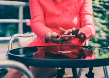 Hands of a young caucasian woman in a pink shirt with a gentle manicure scan the menu on a glass table using a mobile phone poking at it with a finger, sitting at a table in an outdoors cafe, side