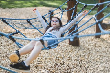 Caucasian girl eight years old with long flowing brown hair in glasses, a gray t-shirt and shorts lying down rides on a rope swing in the park at the playground, side view close-up with selective focus. PARKS REC concept, happy childhood, child picni