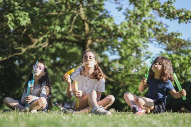 Portrait of three beautiful caucasian girl sisters blowing soap bubbles while sitting in the park on the playground of a flower meadow in backlight, close-up side view with selective focus. The